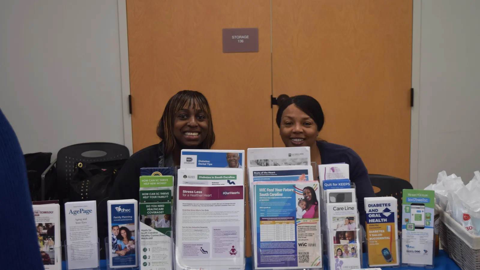 Two women smiling behind a table with health and wellness informational brochures and pamphlets in an indoor setting.