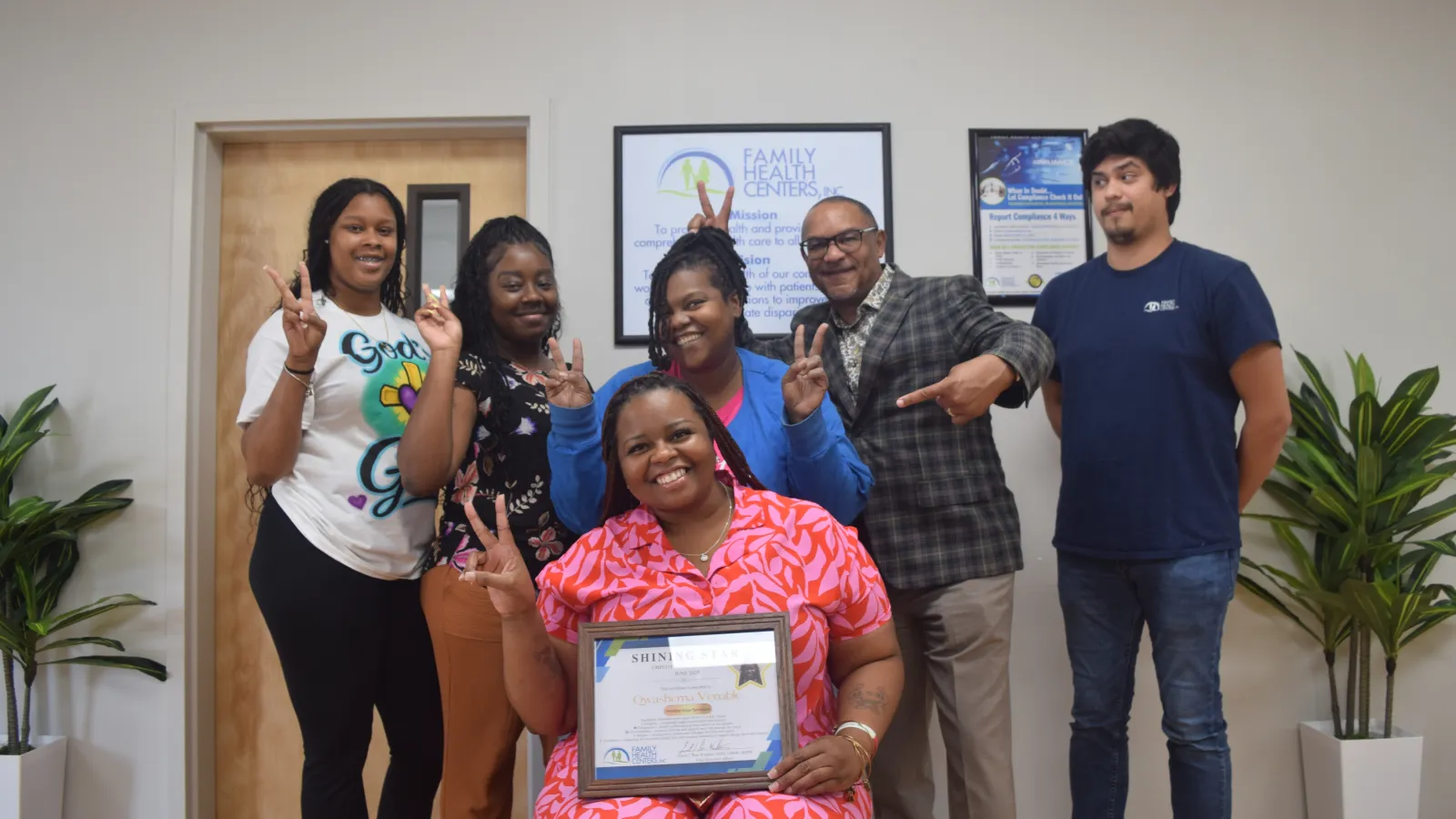 Group of six diverse people smiling celebrating with peace signs, one holding a framed certificate indoors.
