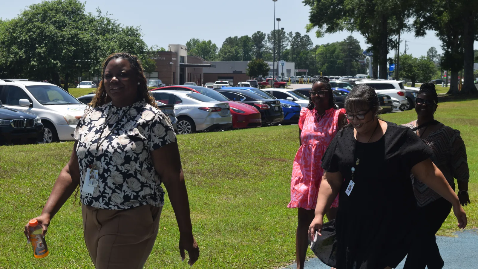 Four women walking outdoors on a sunny day near a parking lot filled with cars and green trees.