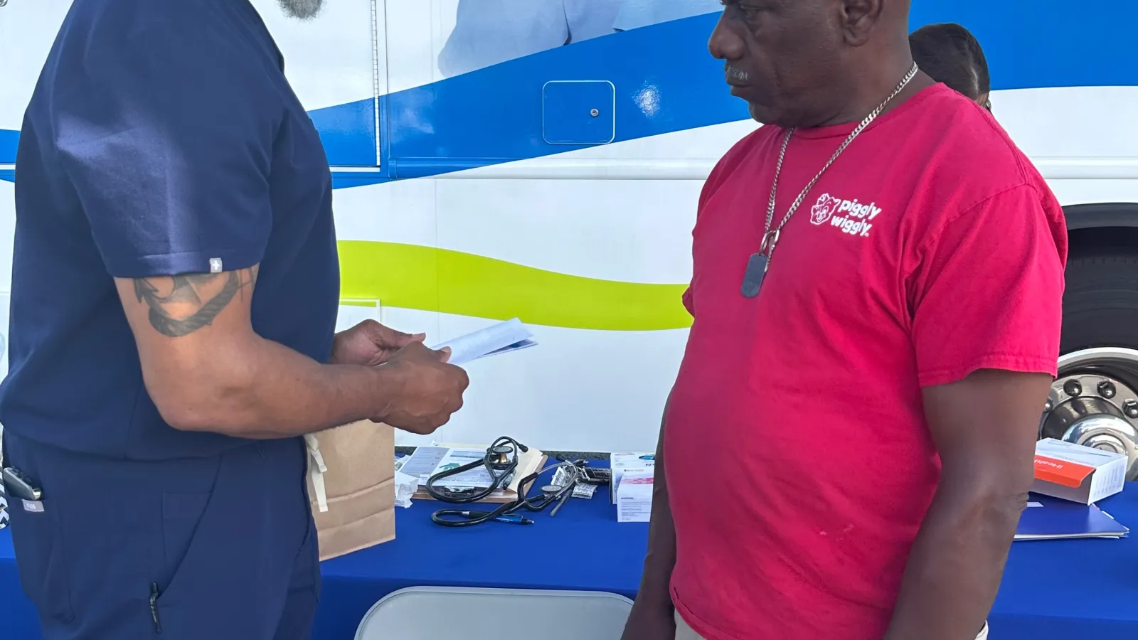 Healthcare professional consulting a man in red shirt at an outdoor medical outreach or mobile clinic booth.