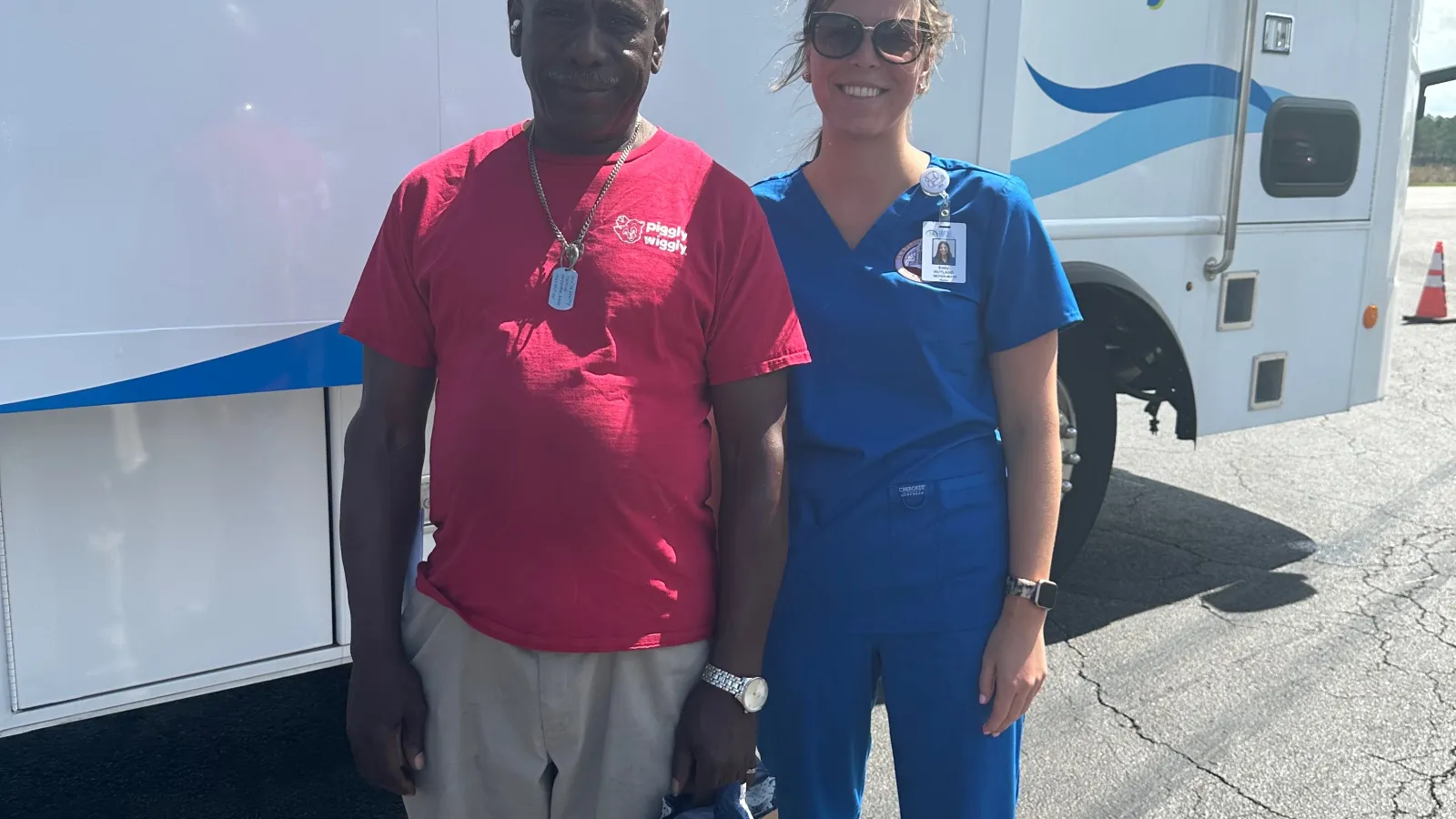 Man in red shirt and woman in blue scrubs standing together in front of health center vehicle with health message.