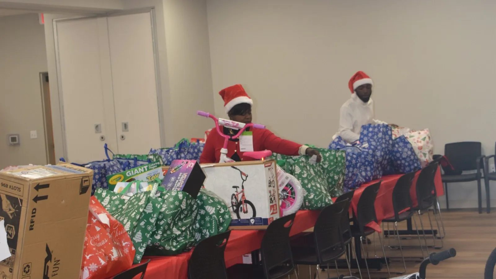 Volunteers wearing Santa hats packing Christmas gifts and bicycles in a festive room with chairs and tables.