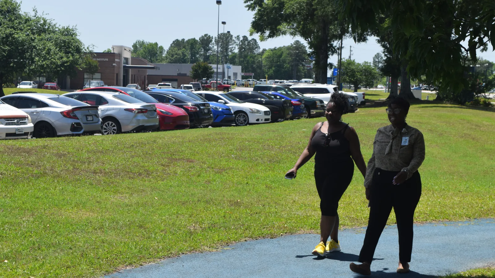 Two women walking on a blue paved path beside a grassy area next to a parking lot with multiple parked cars.