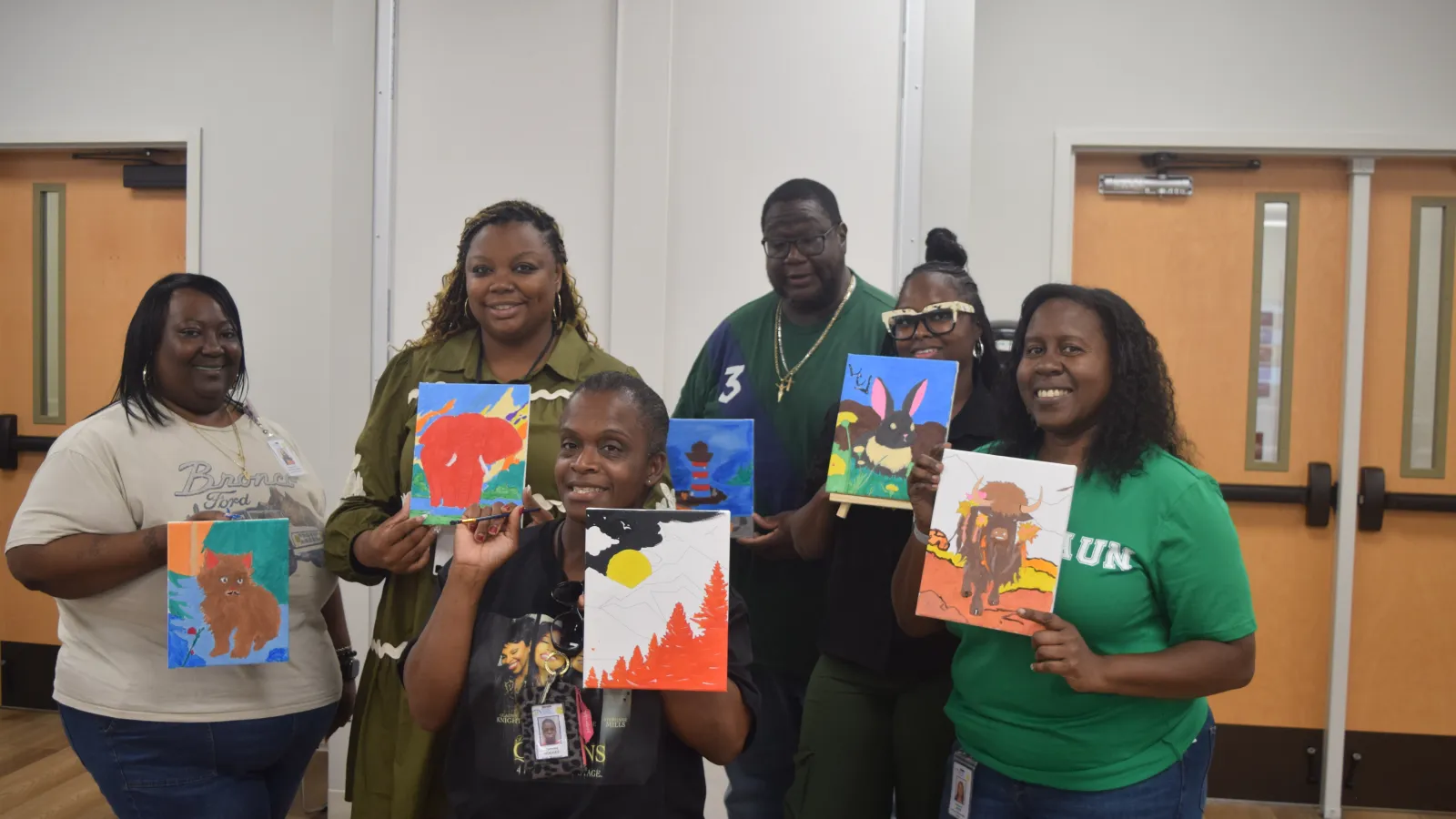 Group of six adults smiling and displaying colorful animal paintings in a classroom setting.