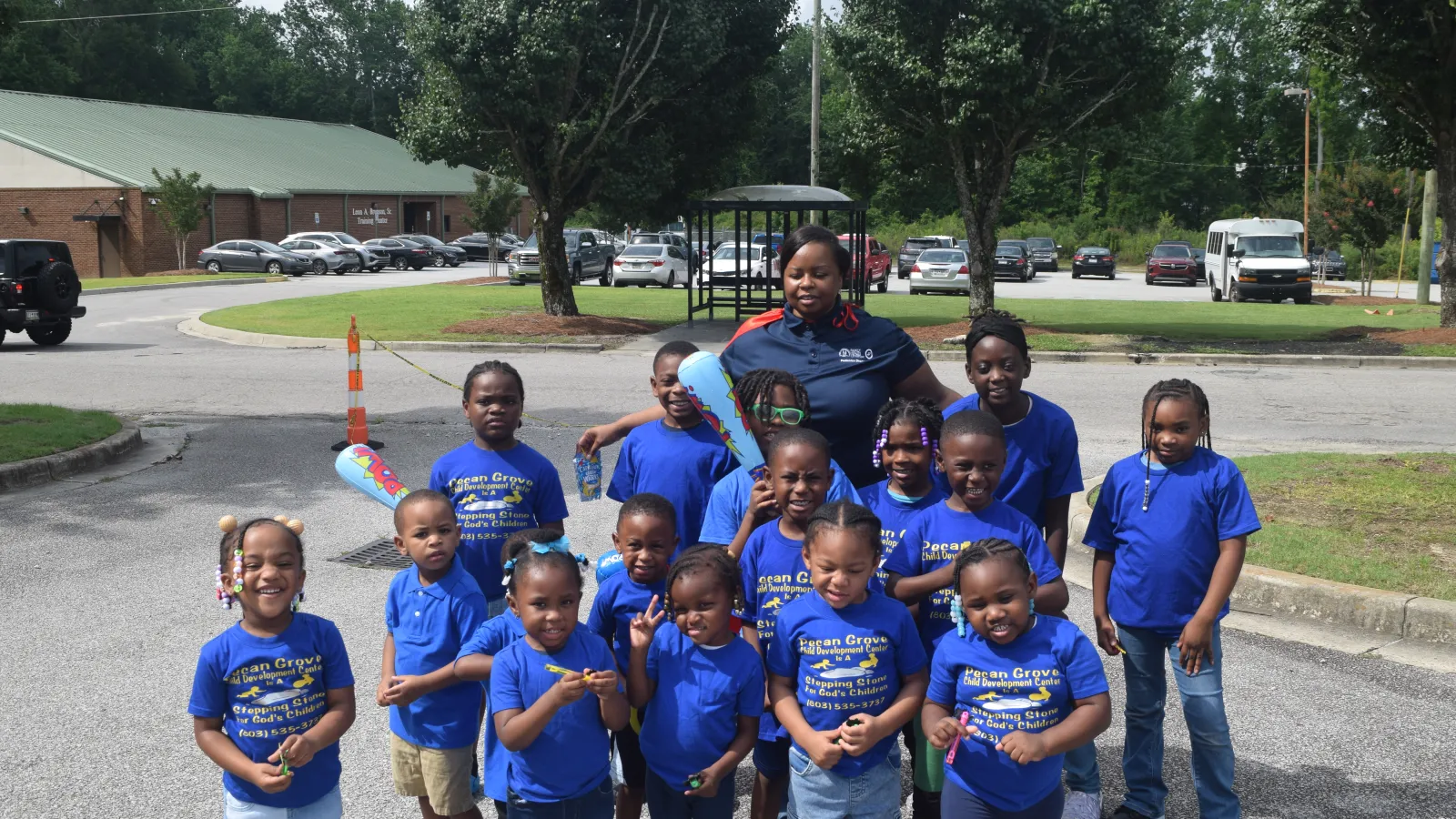 Group of smiling children and a teacher outside wearing matching blue shirts in a parking lot on a sunny day