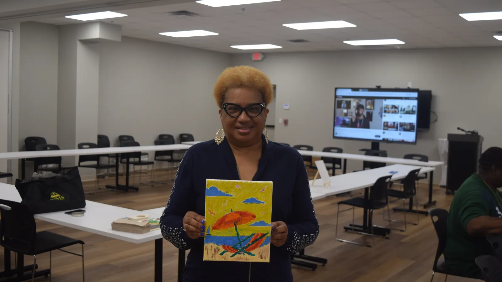 Woman with glasses holding a colorful beach painting in a modern conference room with tables and chairs.