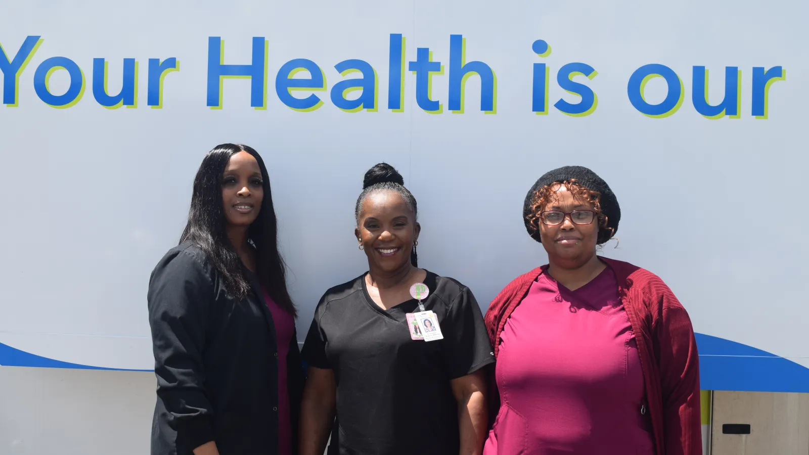 Three smiling healthcare workers posing in front of a sign with the phrase Your Health is Our.