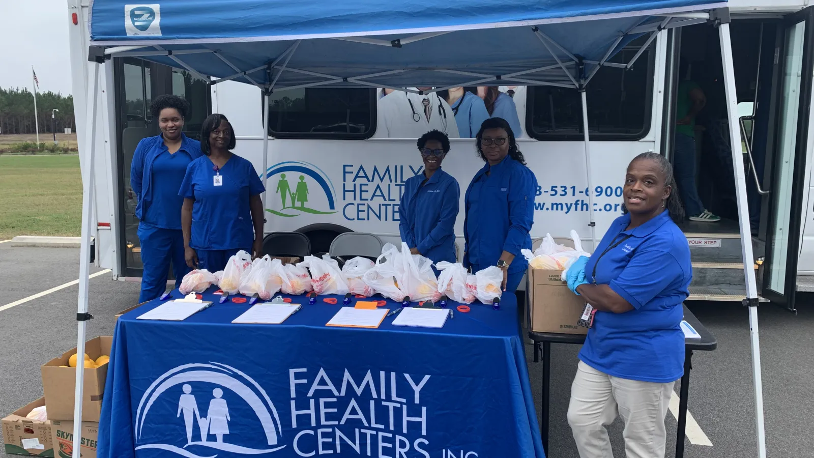 Volunteers in blue uniforms stand behind a Family Health Centers tent with bags of supplies on the table outdoors.