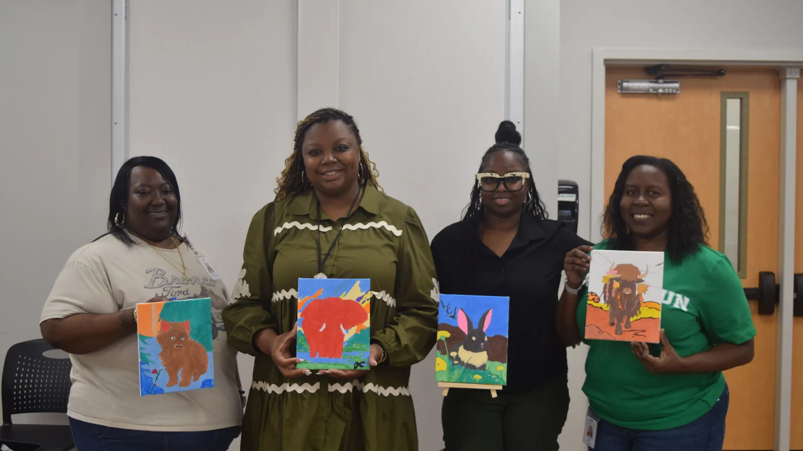 Four women smiling and holding colorful animal paintings in a bright indoor room.