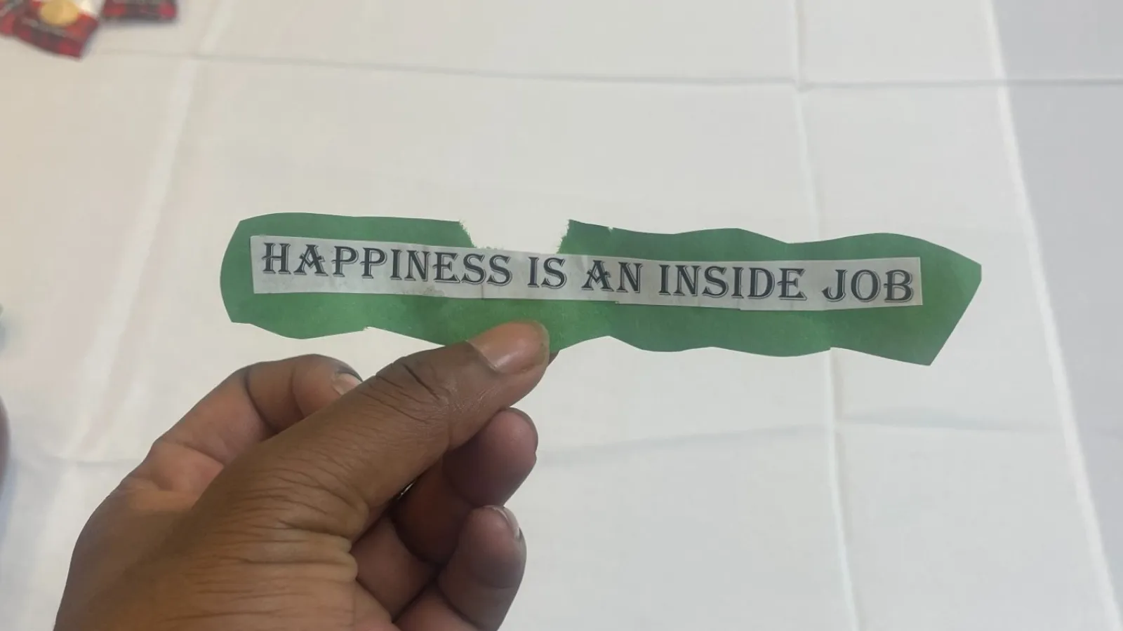 Hand holding a torn paper strip with the phrase Happiness is an inside job on a white table with items scattered.