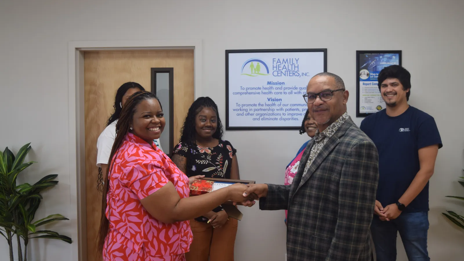 Group of diverse professionals shaking hands and smiling inside Family Health Centers office with mission statement on wall.