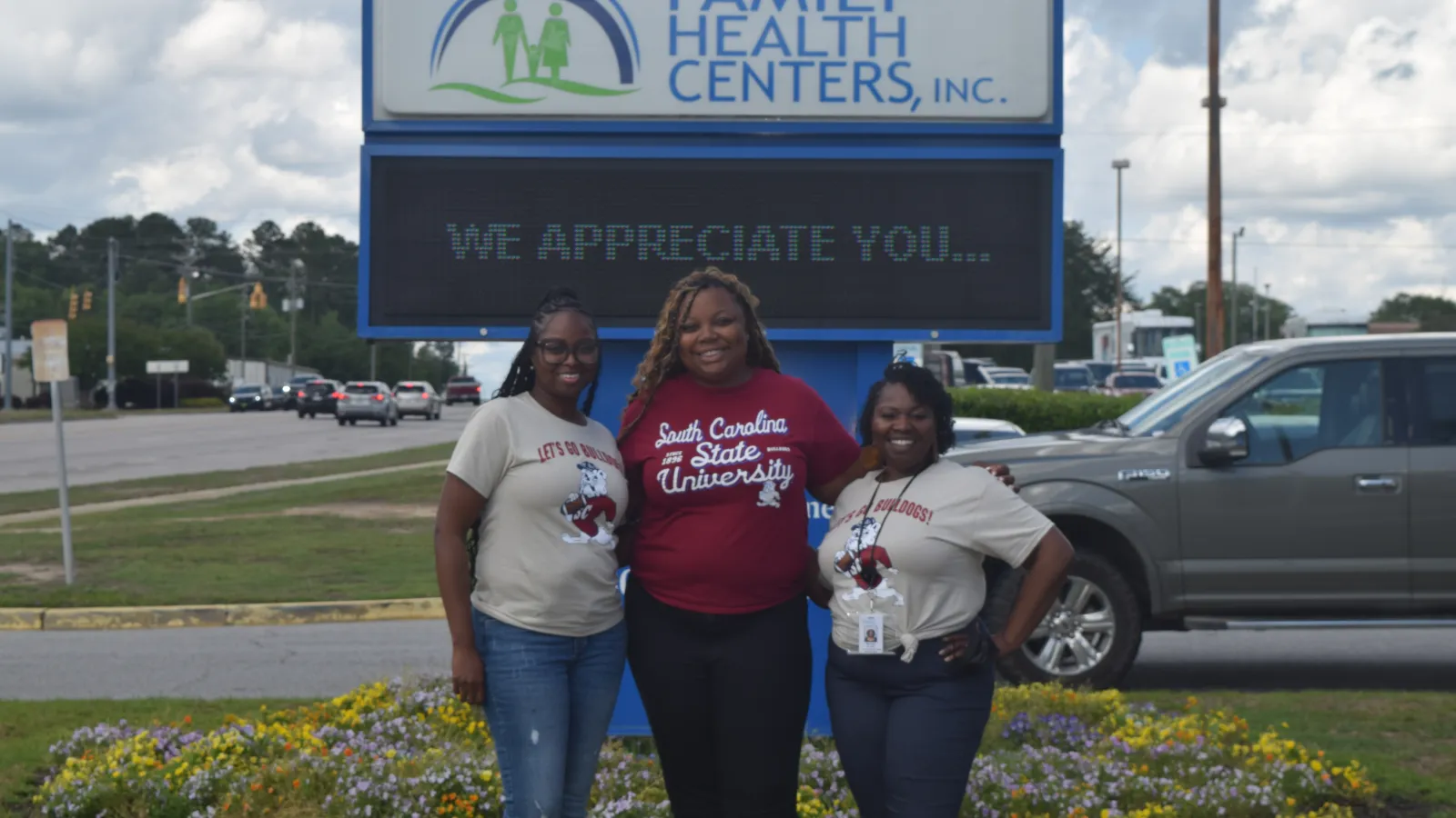Three smiling women standing in front of Family Health Centers sign that reads We Appreciate You outdoors