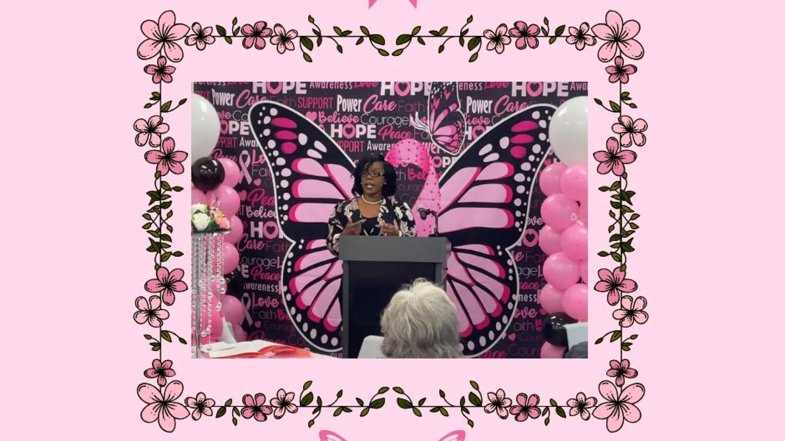 Speaker at podium with large pink butterfly wings and breast cancer awareness decorations.
