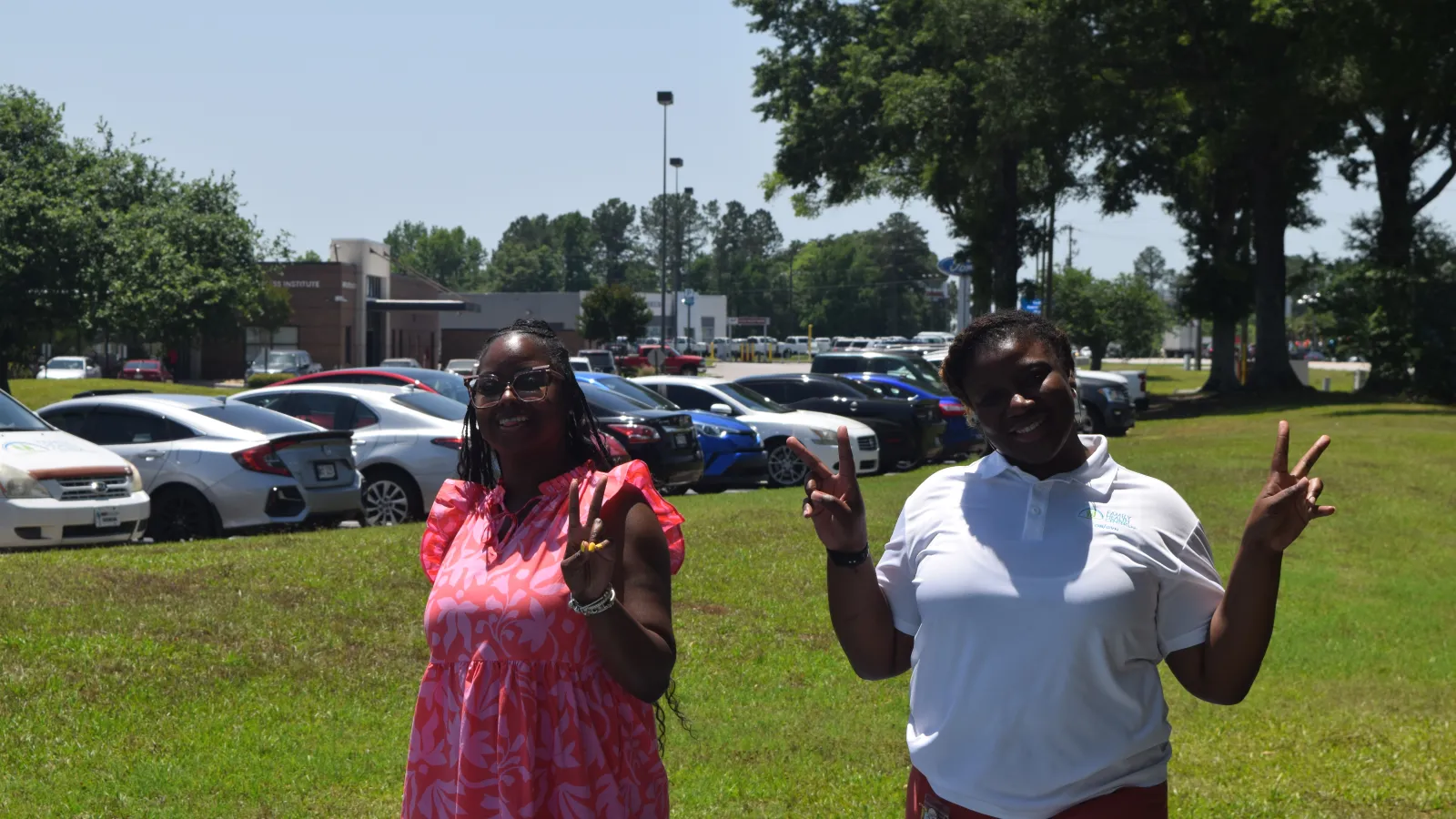 Two women smiling and posing with peace signs outdoors near a parking lot on a sunny day