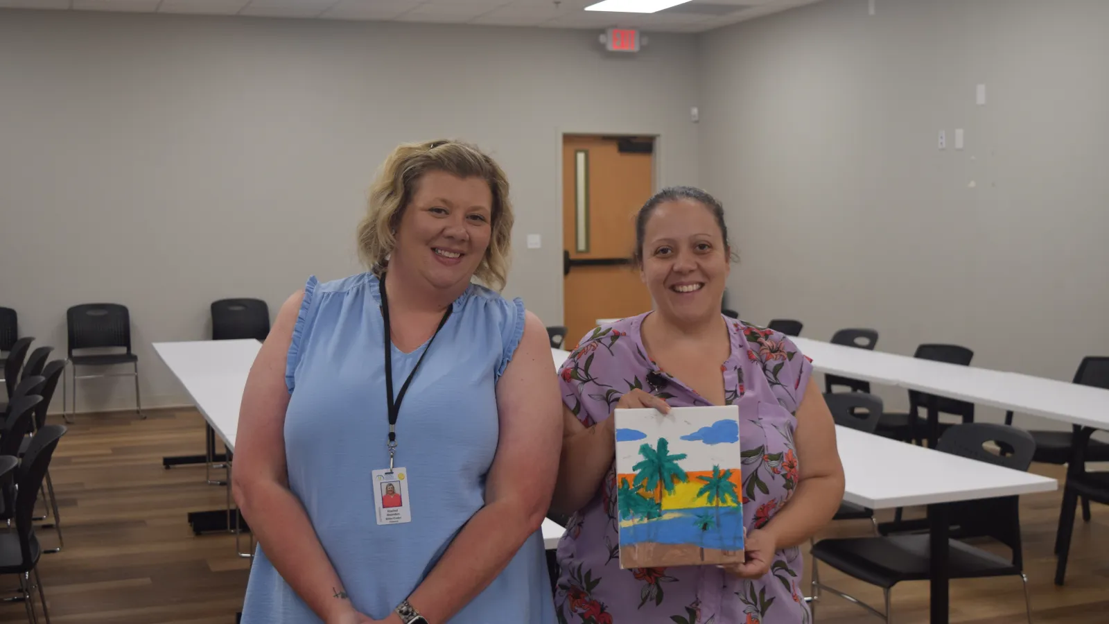 Two women smiling in a meeting room, one holding a colorful palm tree painting, surrounded by tables and chairs.