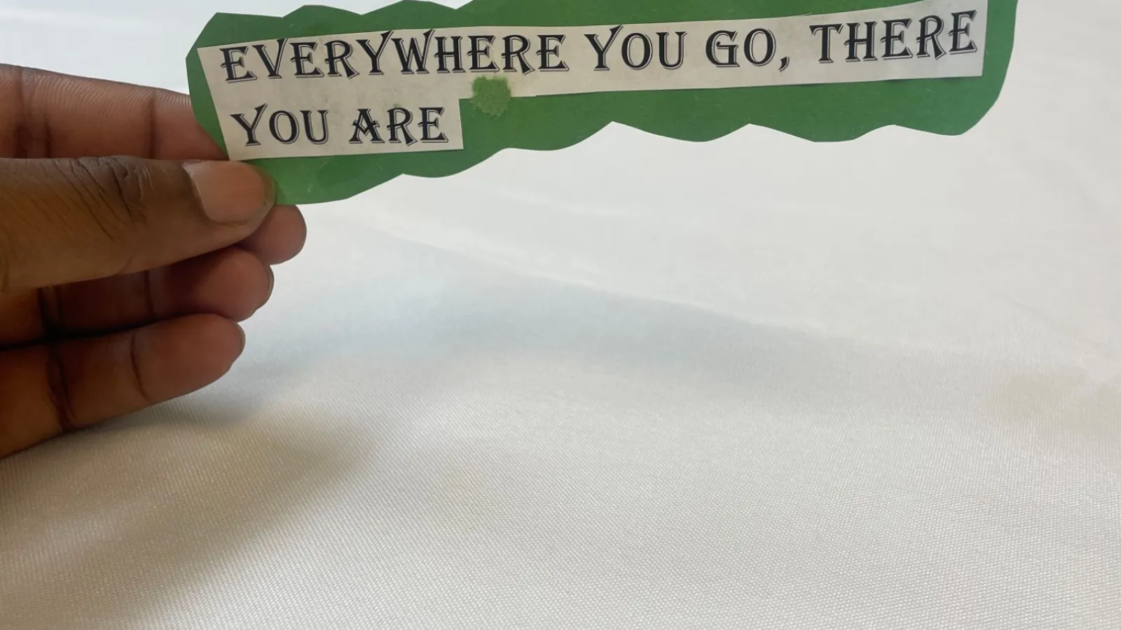 Hand holding a green and white cutout with the text Everywhere you go, there you are on a white fabric background.