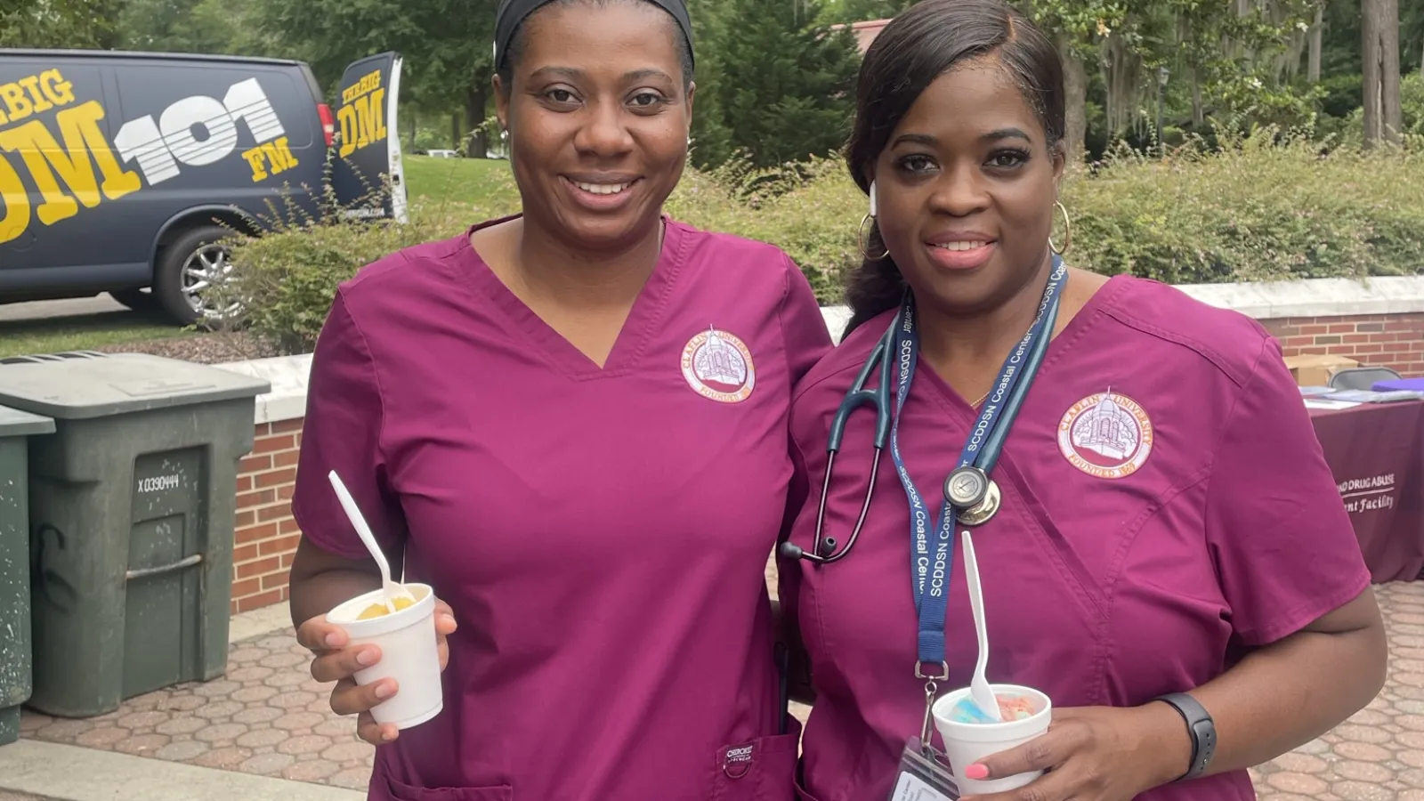 Two smiling nurses in matching purple scrubs holding cups with spoons outdoors on a sunny day.