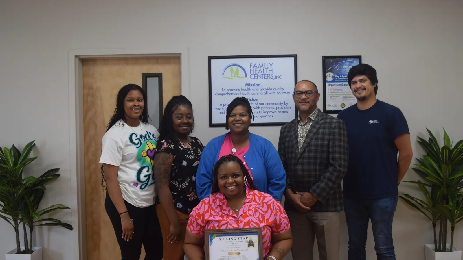 Group of six diverse adults smiling indoors, one holding a framed certificate with plants and signage behind them