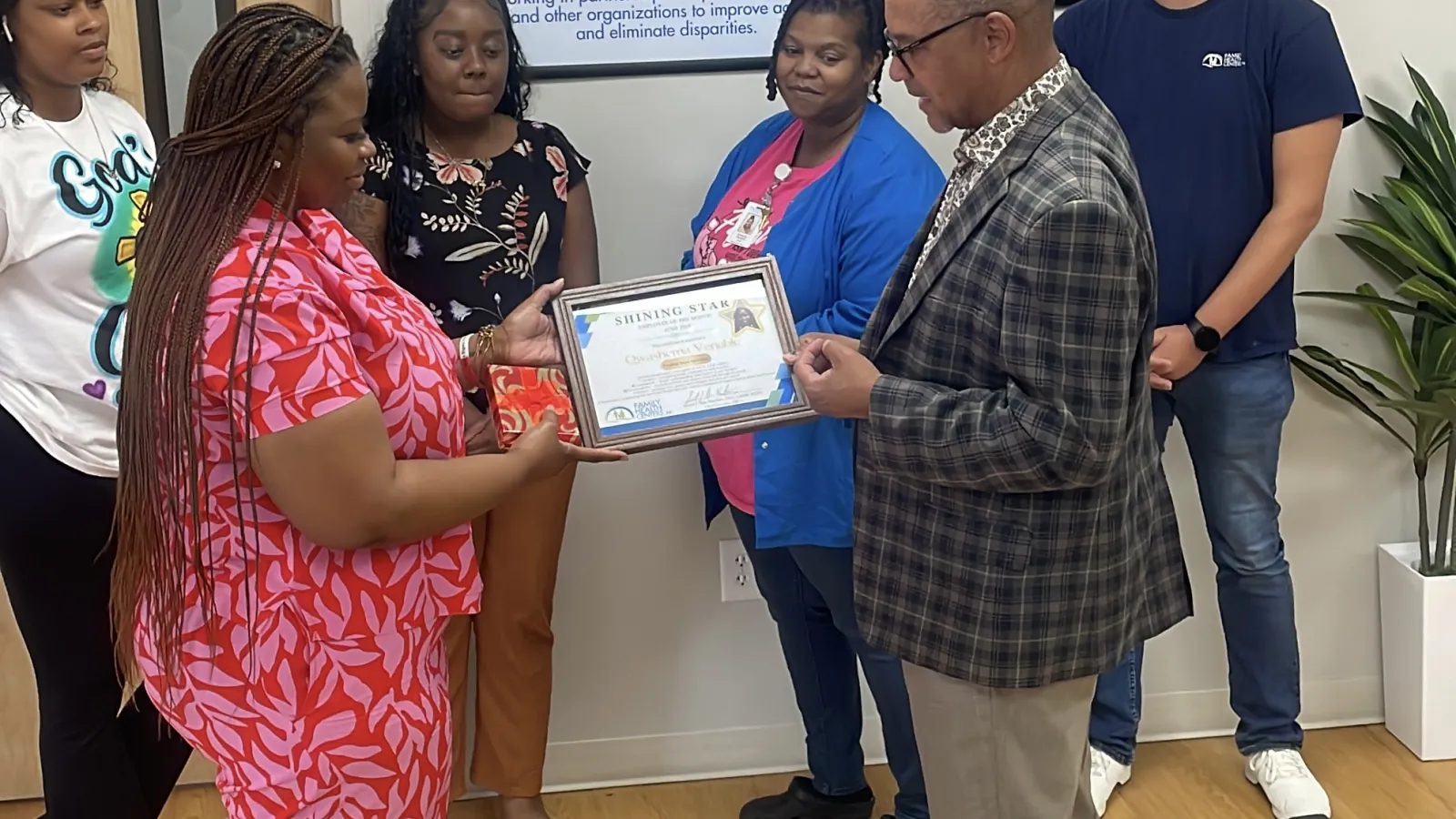 Group of six people in an office setting presenting and receiving a framed certificate award near Family Health Centers sign.