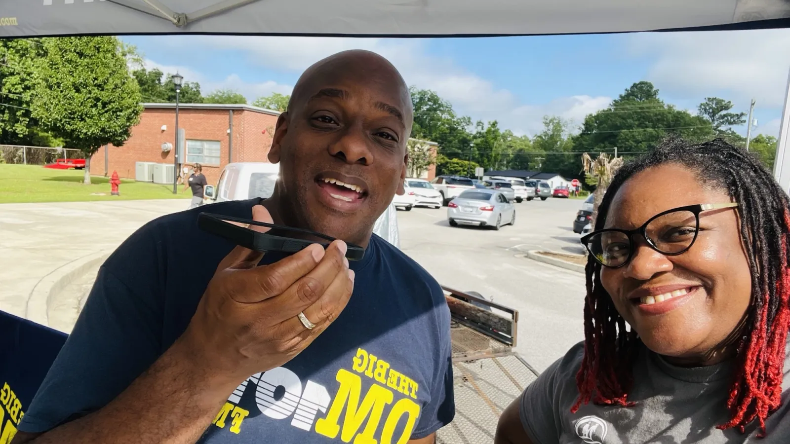 Two smiling people under a tent with THEBIGDM.COM branding on shirts and canopy on a sunny day