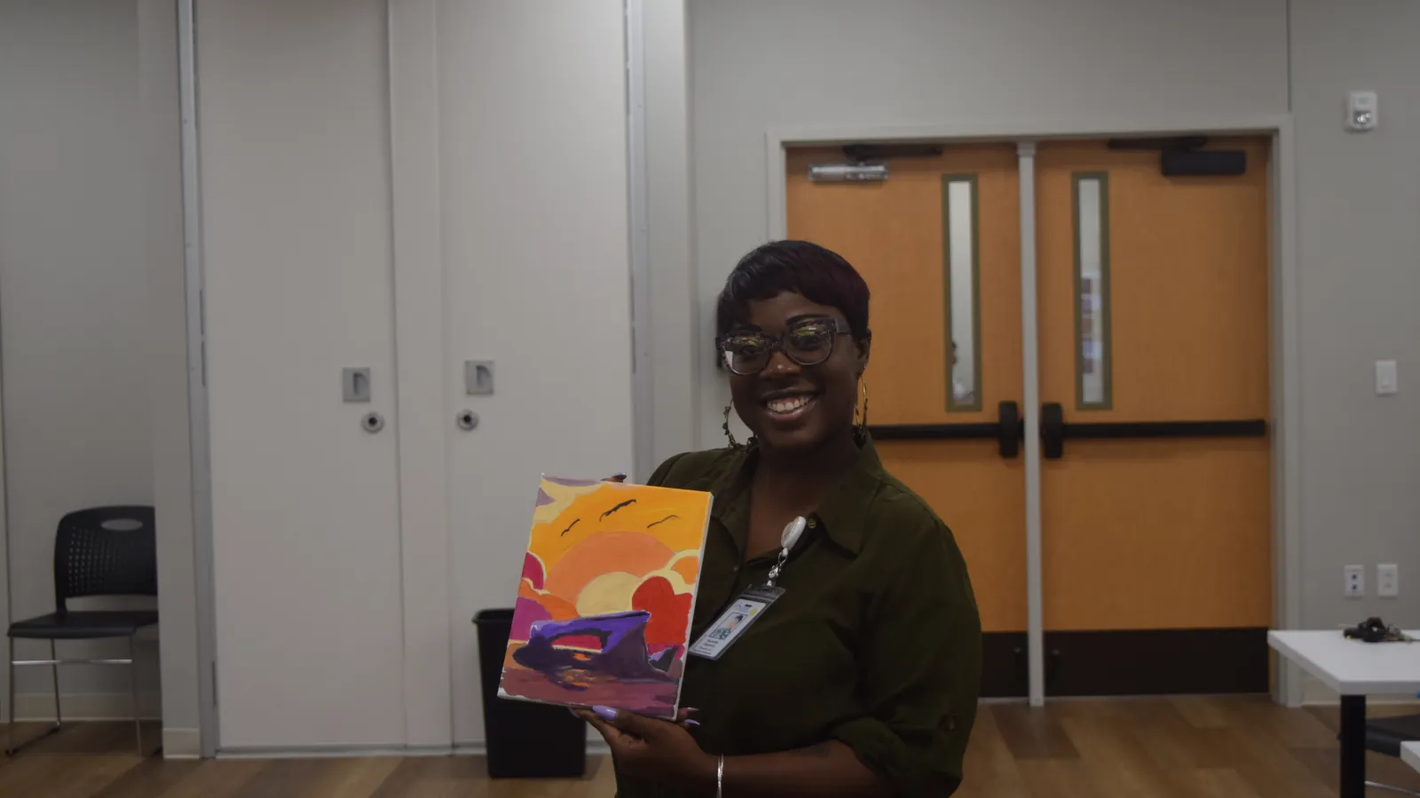 Smiling woman holding a colorful sunset painting inside a room with wooden floors and closed doors.