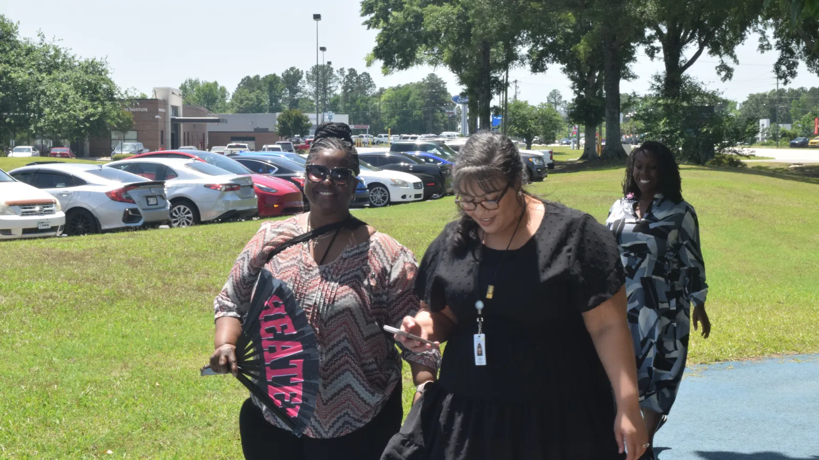 Three women walking outdoors on a sunny day near a parking lot, smiling and enjoying the moment.