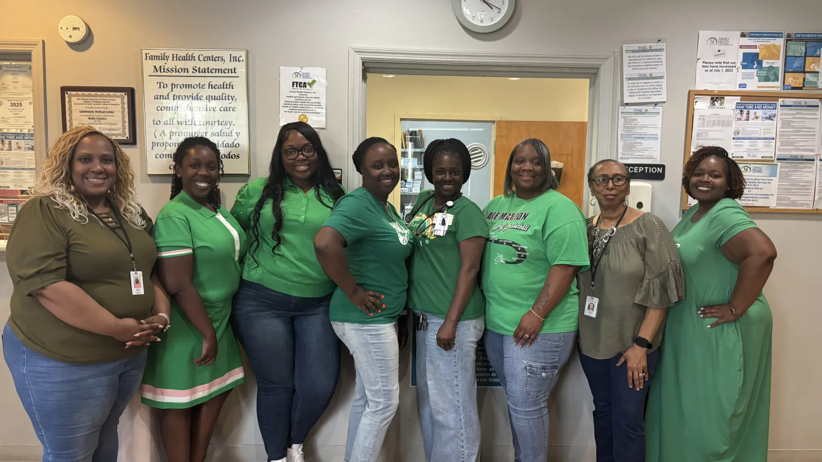 Group of eight smiling women in green outfits standing indoors at a community health center.