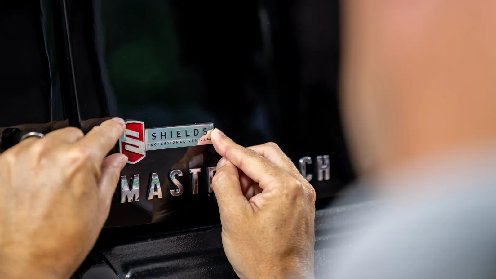 Person applying a Shields Professional Vehicles badge decal to a black vehicle near the MasterTech emblem.