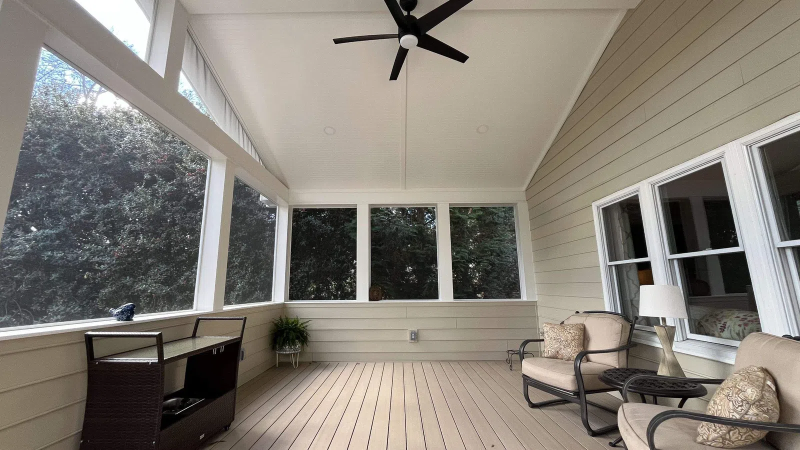 Spacious screened porch with beige wood flooring, two cushioned chairs, black ceiling fan, and natural light.