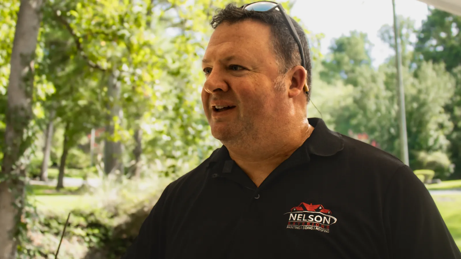 Man in black Nelson Painting polo shirt stands outdoors with trees and sunlight in the background.
