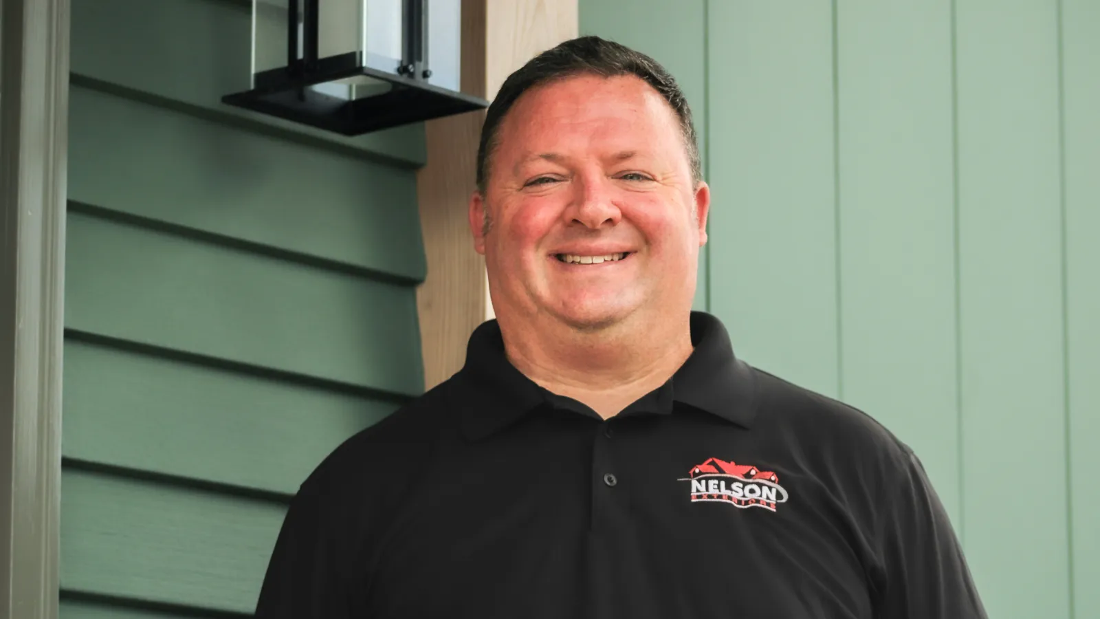 Scott Nelson, President of Nelson Exteriors, wearing black polo stands outside a green building under a black porch light.