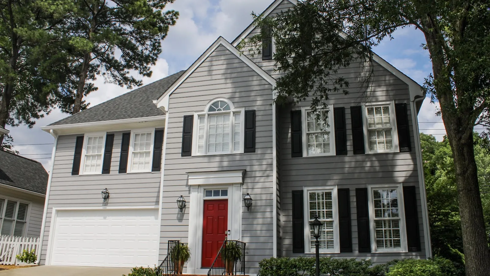 Two-story gray house with white trim, black shutters, red front door, garage, and surrounding greenery under blue sky.