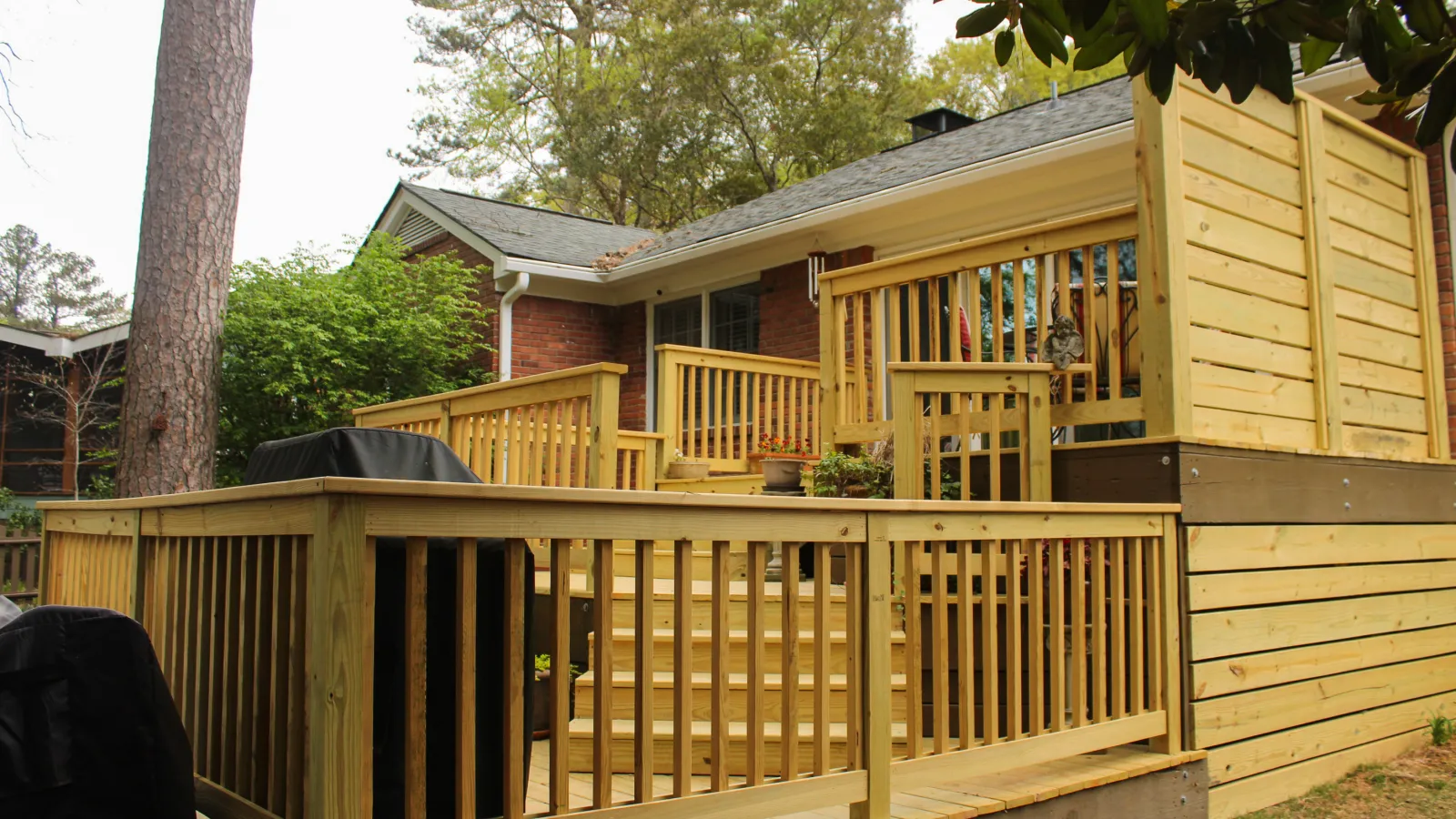Newly built wooden deck with railings and stairs attached to a suburban brick house surrounded by trees.