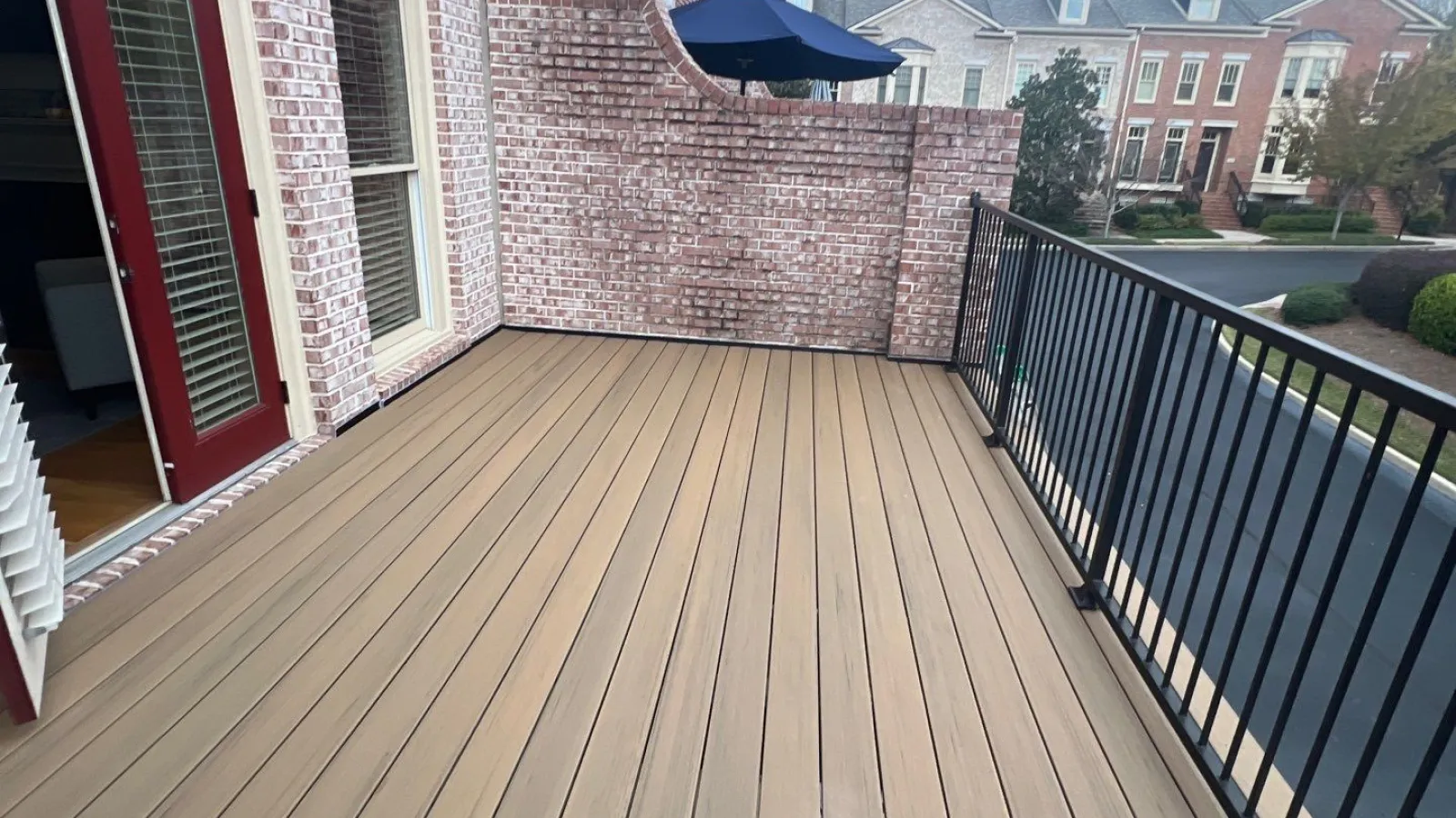 Empty balcony with tan wood decking, brick wall, black metal railing, and cloudy sky in suburban neighborhood.
