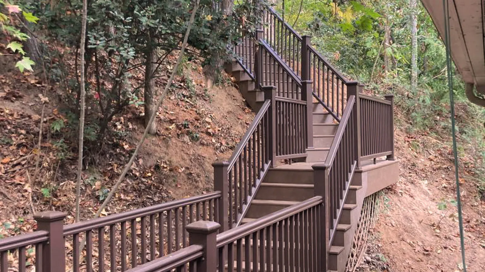 Brown wooden outdoor stairs with railings ascending through a wooded area with autumn leaves on the ground.