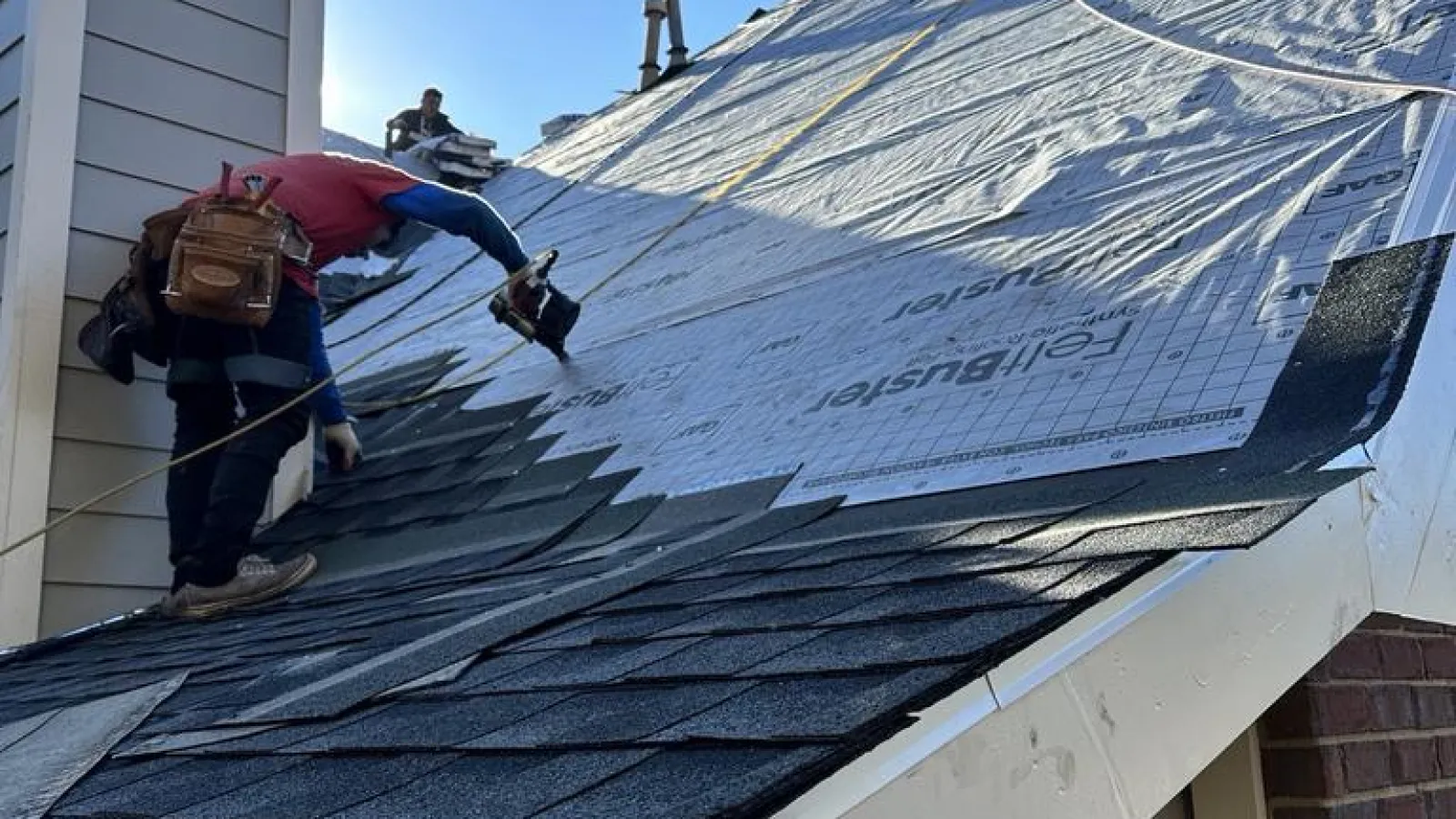 Roofer installing shingles on a sloped roof under clear blue sky during daytime construction work