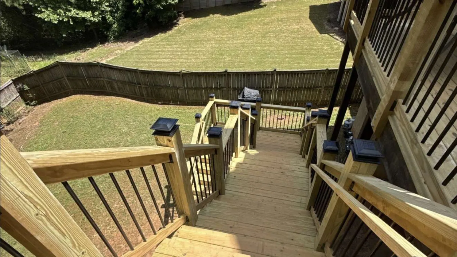 Wooden outdoor staircase with black metal railings leading down to a fenced backyard with grass and trees.