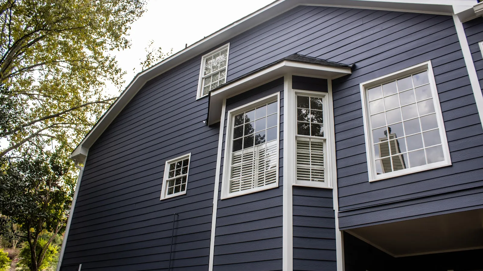 Close-up of green shingle siding on a house with a window and bare tree branches in the background.