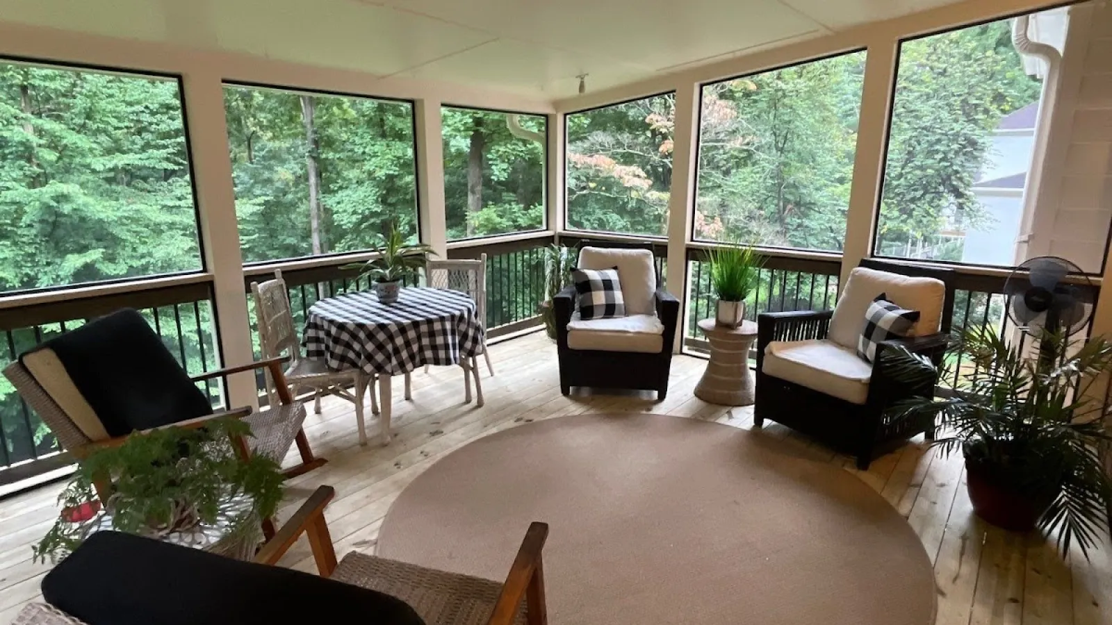 Cozy screened-in porch with wooden floors, wicker rocking chairs, round rug, checkered tablecloth, and green plants.