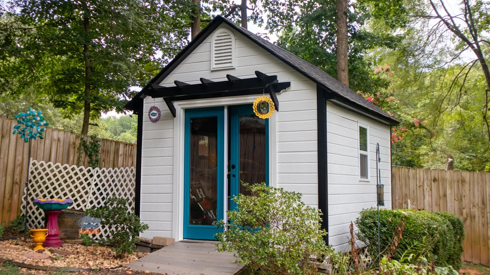 Small white garden shed with blue double doors and black trim surrounded by greenery and wooden fence