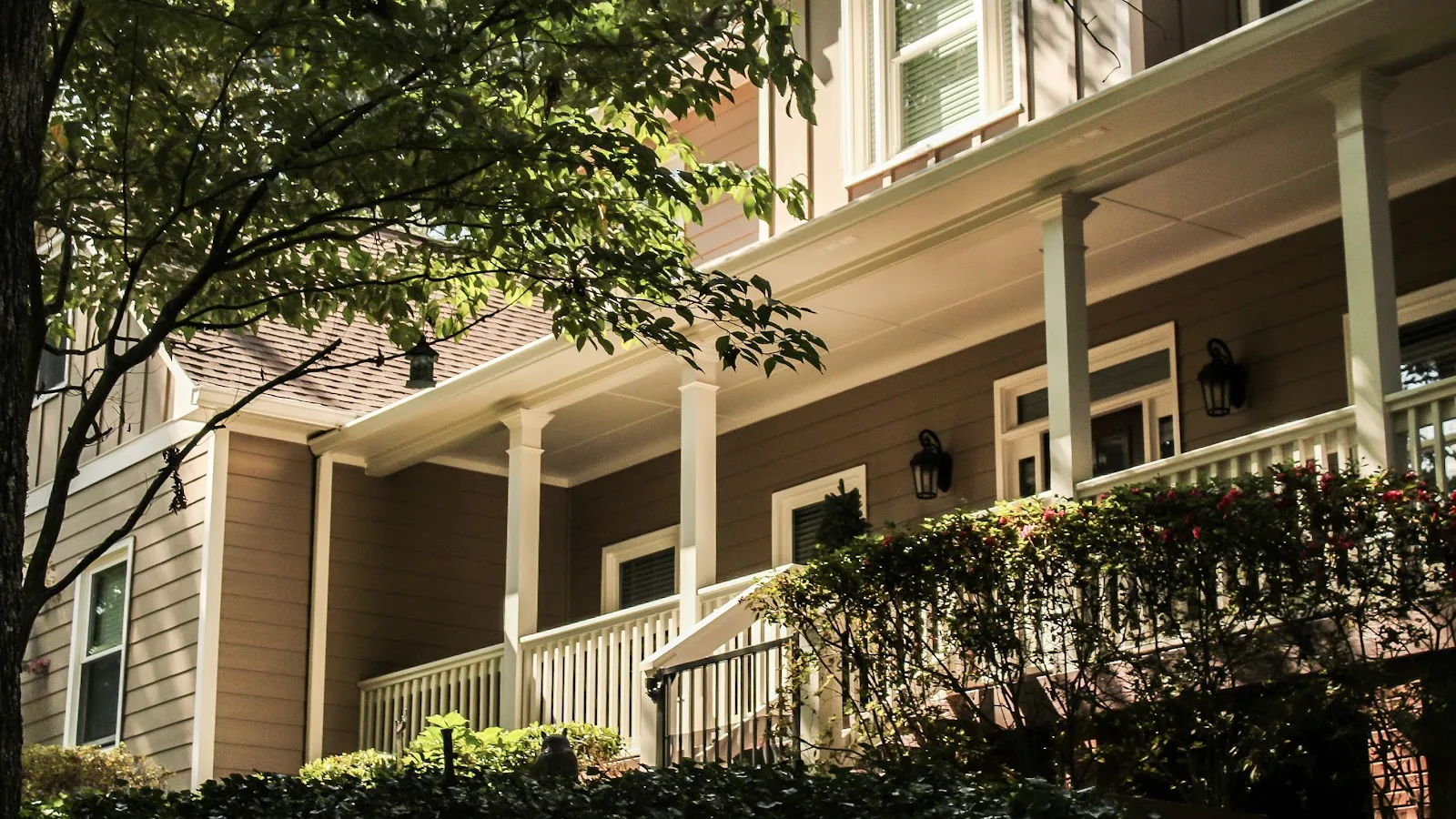Suburban home porch with white railings, beige siding, and lush green trees and bushes under sunlight.