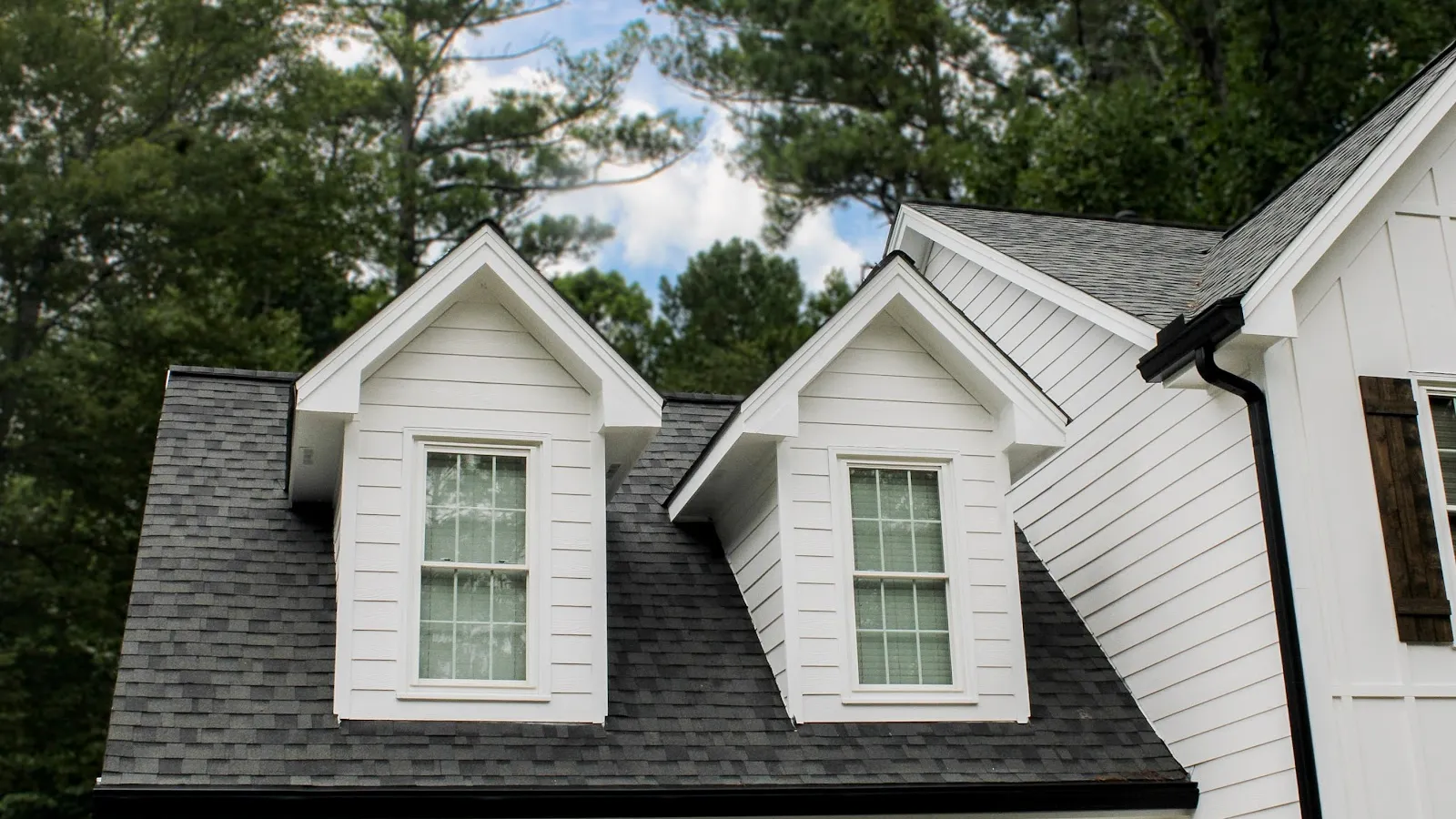 White house dormer windows with black roof shingles surrounded by green trees under blue sky.