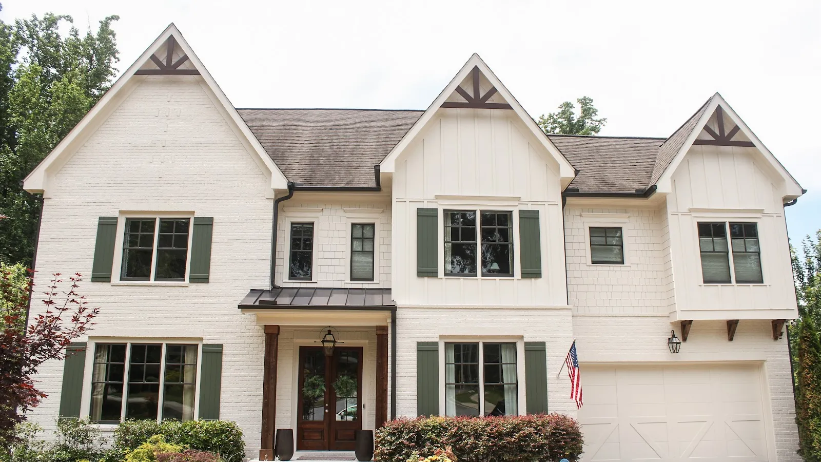 Two-story white suburban house with dark green shutters, a front porch, and an attached garage with American flag.