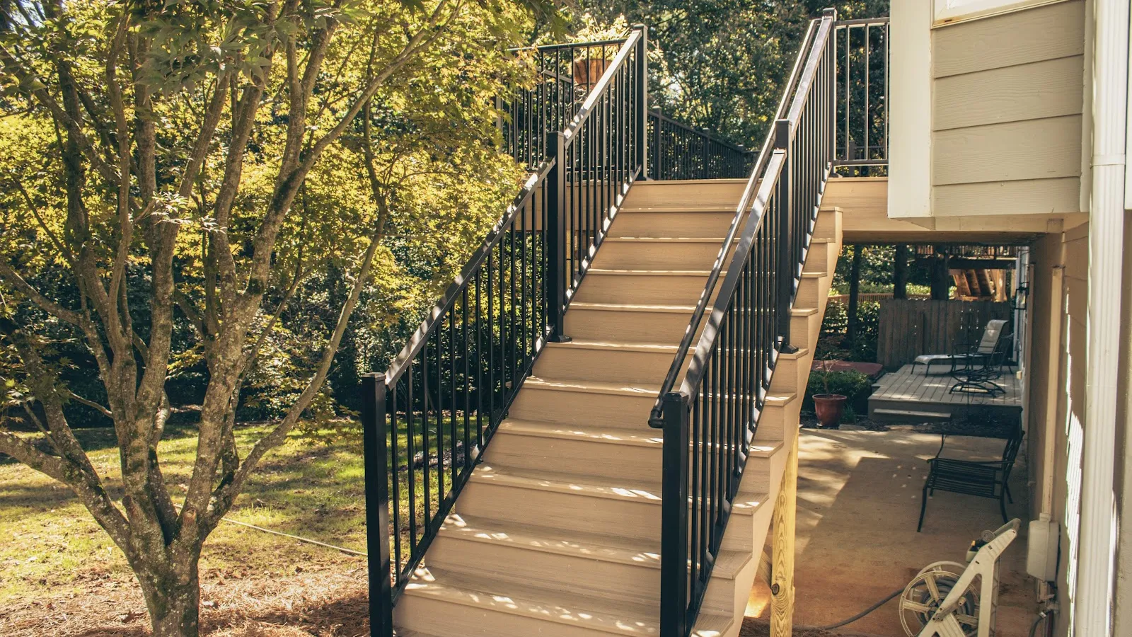 Outdoor wooden stairs with black metal railings leading to a house deck surrounded by trees and garden.