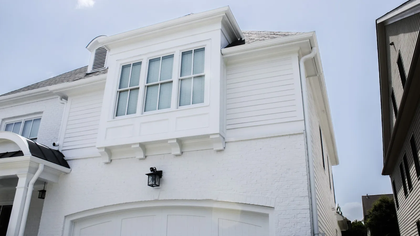 White house dormer windows with black roof shingles surrounded by green trees under blue sky.