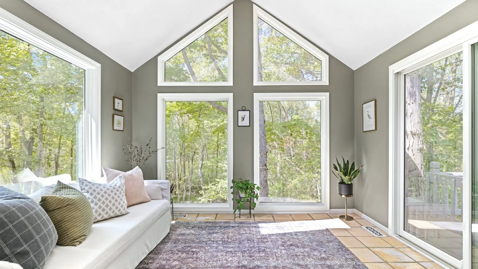 Bright living room with large windows, white sofa, assorted pillows, indoor plants, and forest view outside.