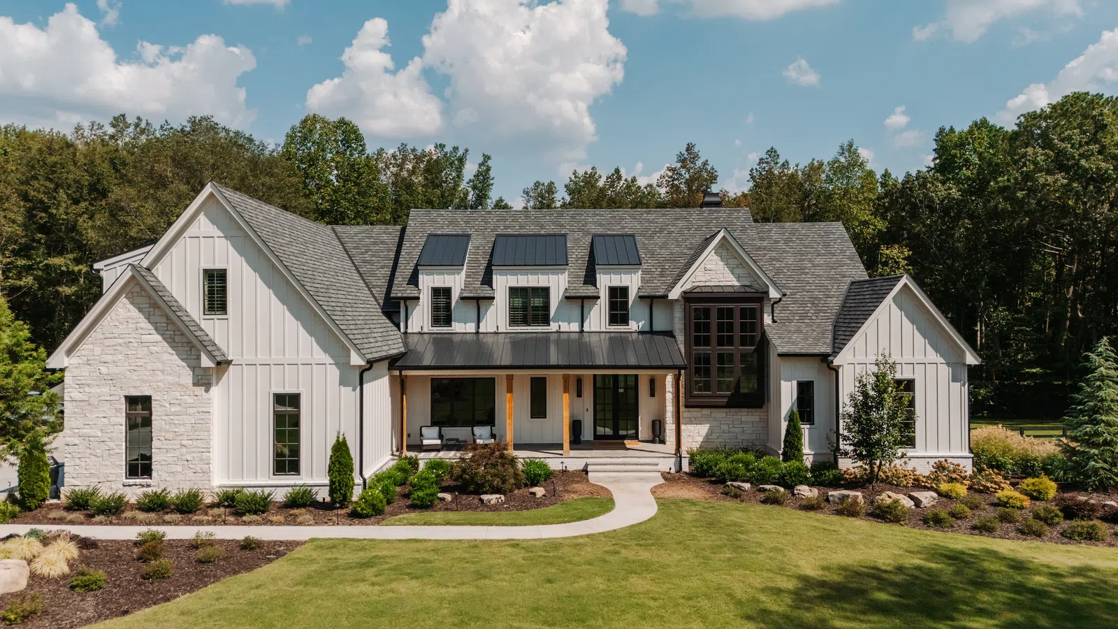 Modern farmhouse with white siding, stone accents, solar panels, and landscaped front yard under a blue sky.