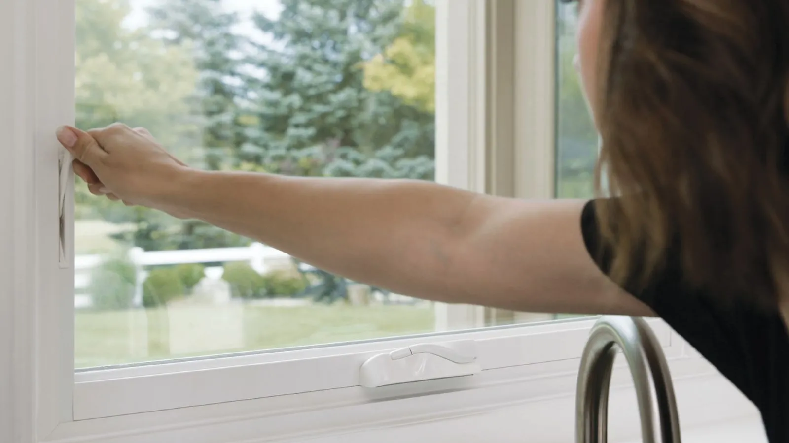 Person opening a white framed window with a view of green trees and lawn outside during daytime.