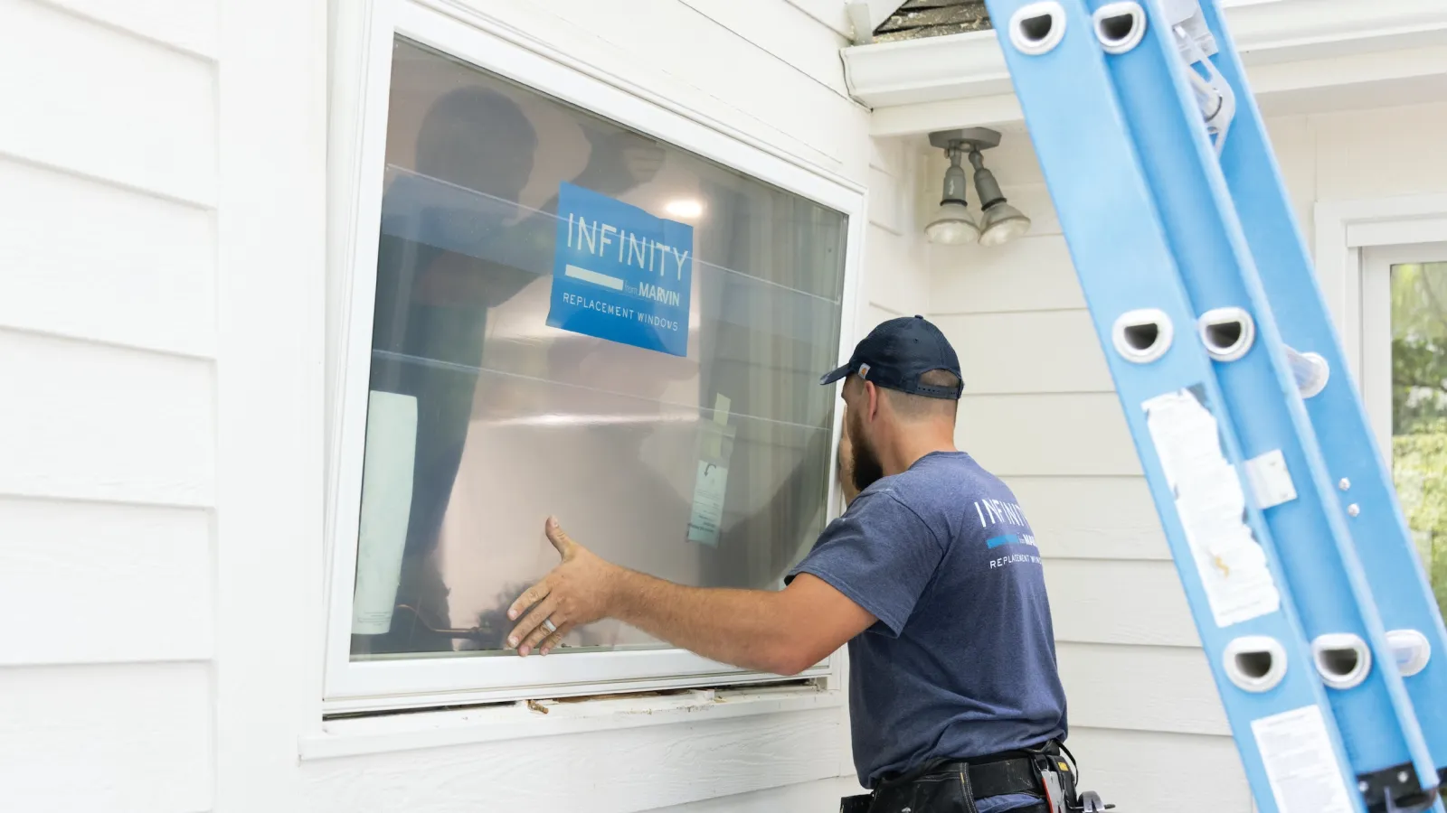 Worker installing Infinity Marvin replacement window on white house exterior with blue ladder.