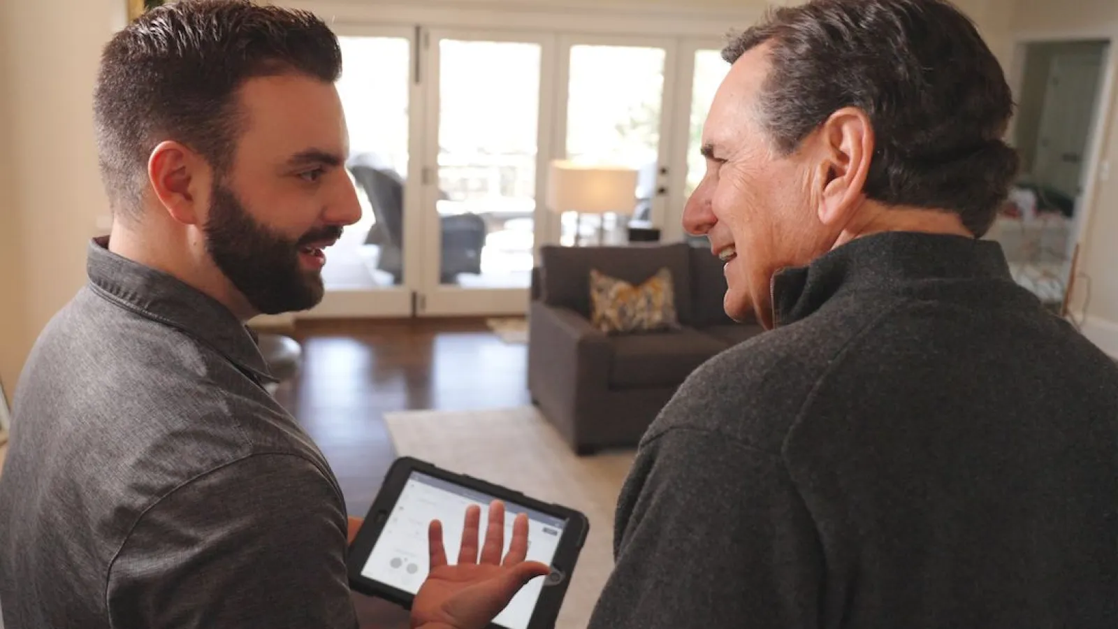 Two men in a living room discussing something on a tablet, with natural light and cozy furniture in the background